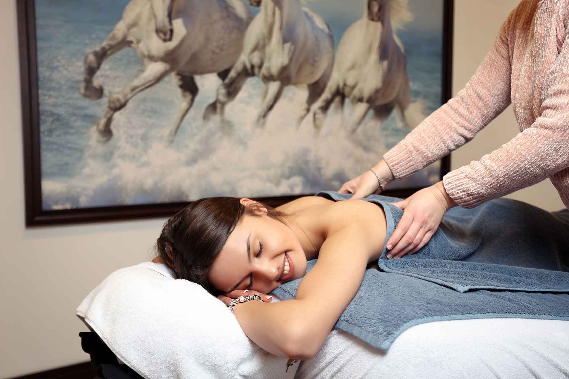 On this picture have a woman on massage bed. Behind her have a picture with three white horses.